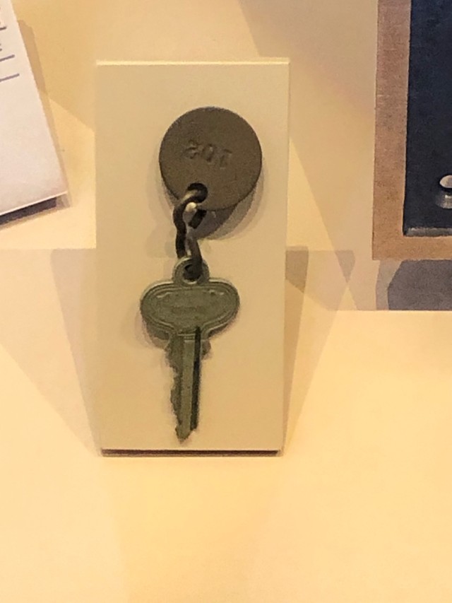 A photograph of a bronze-colored metal key attached to a bronze-colored metal tag featuring the numbers 102. The key and tag are displayed in a case.
